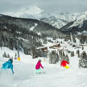 Frontier Ski Named World’s Best Ski Tour Operator to Canada 2025 Three skiers in blue, pink and yellow jackets carve down a snowy slope towards a village nestled beneath towering, forested peaks.