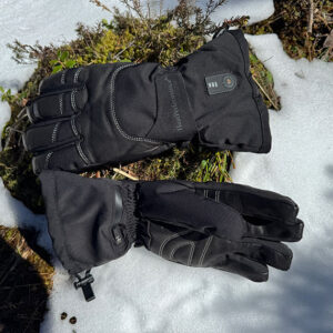 The Hidden Luxury of Warmth A pair of black winter gloves rests on snow-covered heather, featuring a small logo and zip on the wrist.