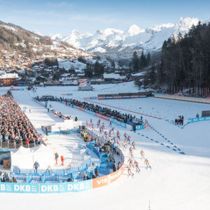 Cross-country skiers race past DKB-branded barriers before large crowds, with snow-capped alpine peaks and a village behind.