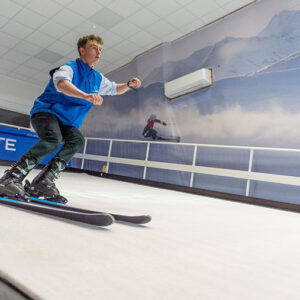 New Indoor Snowsport Training Centre Opens A blonde man in a blue top skis on an indoor slope, with a mountain mural and an "INPISTE" banner behind him.