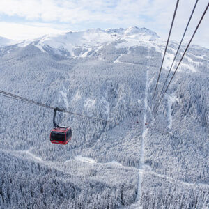 Whistler Blackcomb Celebrates 60 Years A red gondola cabin travels along cables above a vast, snow-covered forest of evergreen trees with mountains in the distance.