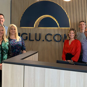 Flight Centre Travel Group acquires Iglu A group of eight people smile beside a reception desk in front of a slatted wall displaying a "CLUBCOM" mountain logo.