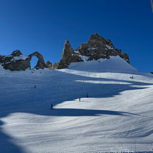 Tignes: before the mountain switches on Skiers descend a sunlit slope towards a jagged rocky peak featuring a natural stone arch, with a chairlift visible on the right under a clear blue sky.