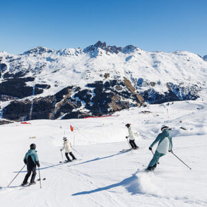 Pick Your Slope: 11 Resorts with Something for Every Skier Four skiers in winter gear descend a wide snow-covered piste, with jagged snow-capped mountains and a valley village visible under a clear blue sky.