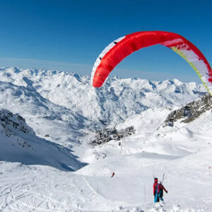A skier walks across a vast snowy mountainside carrying skis, with a large red and white paraglider soaring above the snow-capped peaks under a clear blue sky.