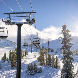 Adventure awaits skiers and boarders at Mammoth Lakes Empty chairlifts ascend a snow-covered mountainside dotted with pine trees beneath a bright sun and blue sky.