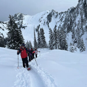 Winter activities in Gstaad & Interlaken Three people in winter gear follow a single-file track through deep snow towards a steep, snow-capped mountain flanked by evergreen trees.