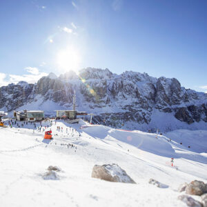 Olympic Torch to pass through Val Gardena Skiers gather on a sunlit snow slope below rugged mountain peaks and a cluster of alpine buildings.