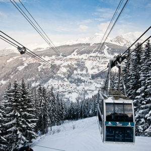 A cable car marked "Les Arcs" descends a snow-covered mountainside towards a ski resort village nestled in the valley below.