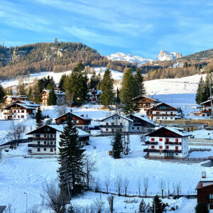 Winter Olympic flame ready to light up Italy A snow-covered alpine village of traditional wooden chalets sits among evergreen trees, backed by forested mountains under a blue sky.