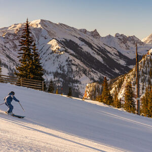 A first-timer’s guide to Aspen Snowmass Skier in a blue jacket carves down a snowy slope, kicking up powder with sunlit mountains, evergreens and a wooden fence behind.
