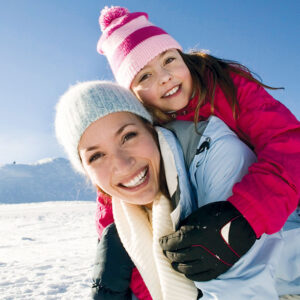 Smiling woman in a white hat carries a young girl in a pink jacket on her back across snow.