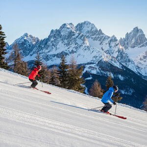 Ski where Olympic champions made history Two skiers in red and blue jackets carve down a groomed piste beneath jagged, snow-covered mountain peaks and scattered evergreen trees.