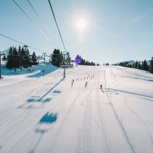 SkiWeekends' new packages for Les Gets special beginners' week Skiers descend a wide, sunlit piste viewed from a chairlift, their shadows stretching across the snow beneath a bright sun.