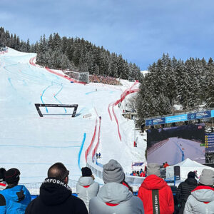Spectators in winter jackets watch a ski race on a snowy mountain course marked with red netting, alongside a large results screen.