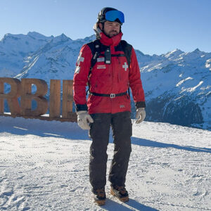 Helly Hansen celebrate International Ski Patrol Day Piste patroller in a red HH jacket and helmet with blue goggles stands on a snowy summit beside a wooden Verbier sign.