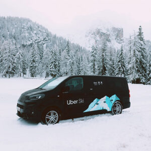 Uber brings new rides to the slopes for the Olympic and Paralympic Winter Games Black Uber Ski van with mountain graphic parked in snow, surrounded by snow-covered pine trees and a misty mountain backdrop.