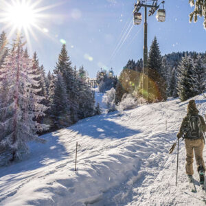 Late Season Skiing in Saint-Gervais Mont Blanc A skier with a backpack and a black dog ascend a sunlit snowy slope beneath a cable car, surrounded by frost-covered pine trees.