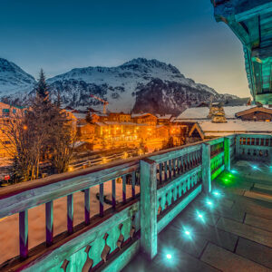 Last-Minute Luxury Escapes to the French Alps A teal-lit wooden balcony overlooks a snow-dusted alpine village and snow-capped mountains at dusk.