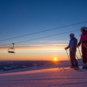 Scandinavia: A winter wonderland for family ski holidays Two skiers in helmets and goggles stand on a snowy slope at sunset, beside empty chairlift cables, with an orange glow on the horizon.