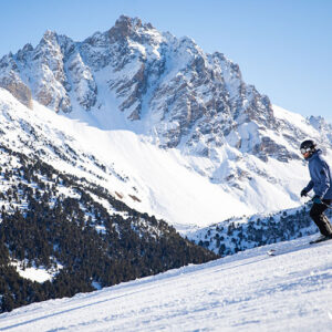 Skier in a blue jacket and helmet descends a groomed slope with a dramatic snow-capped mountain rising behind, marked with the Méribel logo.