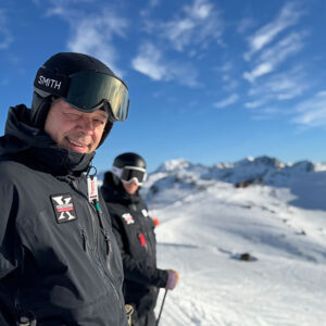 Whistler at 60: One Man Who Helped Build Its Steep Soul Two skiers in black helmets and jackets stand on a snowy mountain ridge, with the lead skier wearing a Smith helmet beneath a bright blue, wispy-clouded sky.
