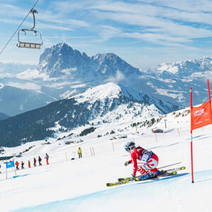 Val Gardena to host longest giant slalom in the world Skier in a red racing suit carving around a red gate on a snowy slope, with chairlifts overhead and rugged snow-capped mountains in the background.
