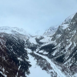 Snow-covered mountain peaks rise above a wide valley with a winding road and scattered trees under a grey, overcast sky.