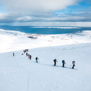 Luxury ski adventures in Iceland and Argentina A line of skiers ascends a snow-covered slope in single file, with a coastal town and fjord visible far below under a partly cloudy sky.