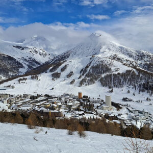 A snowy mountain resort village sits in a valley surrounded by tree-dotted slopes and cloud-capped peaks under a blue sky.