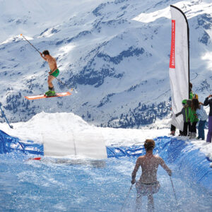 Celebrate the end of season at the Ultime Session A shirtless skier in green shorts launches off a snow ramp into a water pool, with spectators watching and snow-capped mountains rising behind.