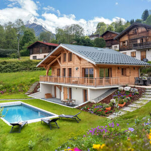 Wooden alpine chalet with balcony and garden swimming pool, set against snow-capped mountains and traditional hillside buildings.